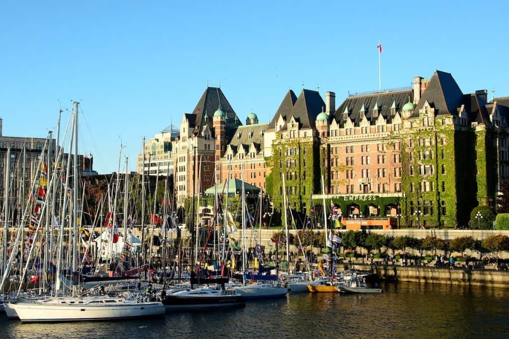 A bustling marina filled with sailboats in front of the iconic ivy-covered Fairmont Empress hotel. The historic architecture, with its green-roofed turrets, stands prominently against a clear blue sky, reflecting the maritime heritage of the city.